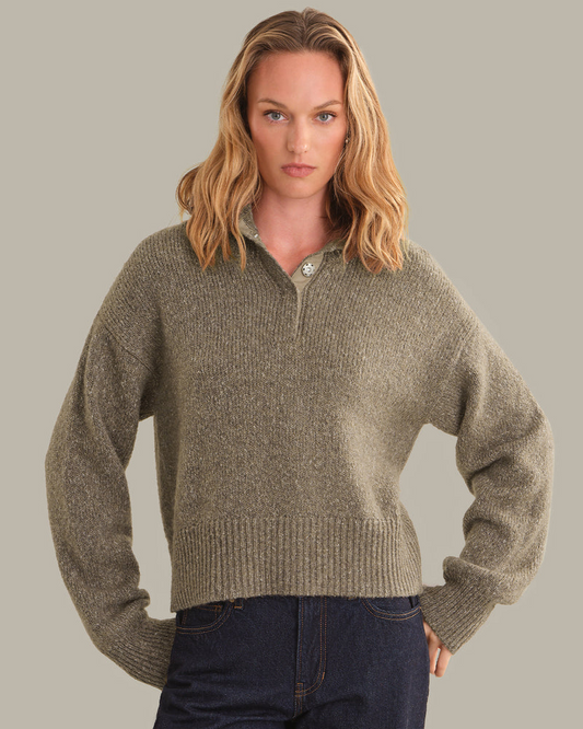 A woman modeling a cropped, ribbed knit half-zip sweater in a heathered taupe-brown color, paired with dark blue jeans, against a solid light gray background.