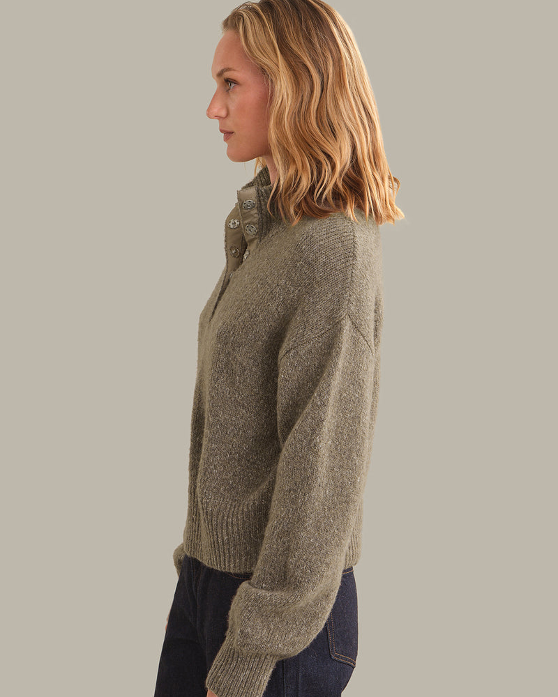 A woman in profile wearing a textured, heathered taupe-brown knit sweater with a high buttoned collar, paired with dark blue jeans, against a solid light gray background.