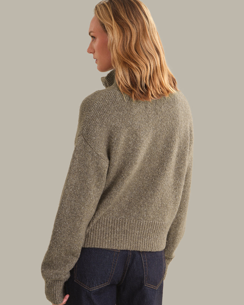 A woman shown from the back wearing a textured, heathered taupe knit sweater and dark blue jeans, set against a solid neutral gray background.
