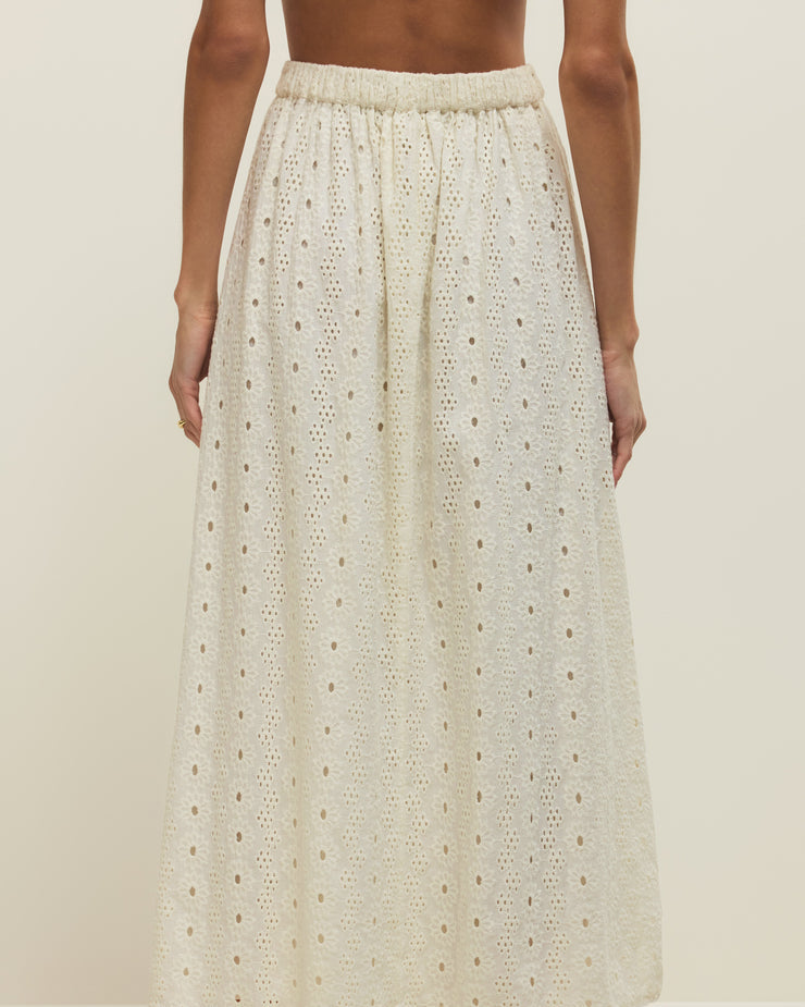 Person wearing a long, light-colored skirt with a subtle pattern on a plain background