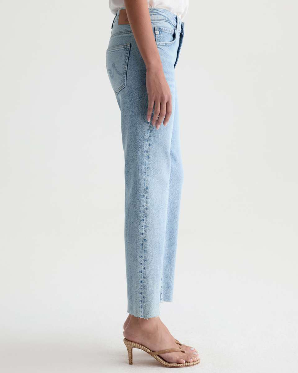 A side profile view of a person wearing light-wash blue cropped jeans with a decorative vertical line of dark studs down the left leg, styled with a white top and beige heeled sandals against a plain white studio background.