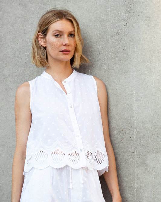Woman wearing a white sleeveless top with lace details against a gray concrete wall.