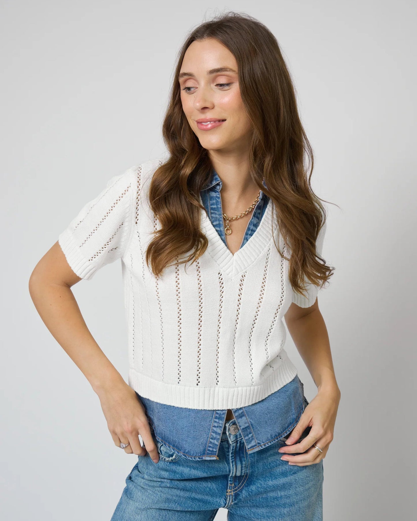 Woman wearing a white sweater over a denim shirt with blue jeans on a plain background