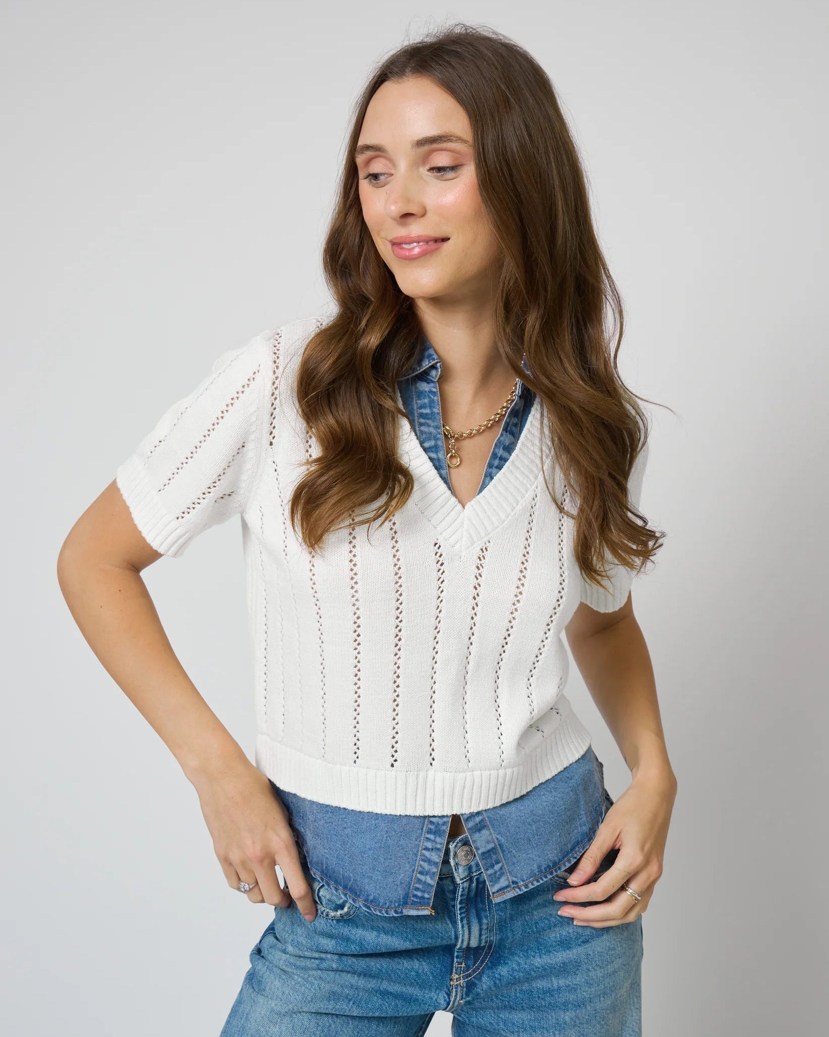 Woman wearing a white sweater over a denim shirt with blue jeans on a plain background