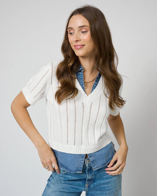 Woman wearing a white sweater over a denim shirt with blue jeans on a plain background
