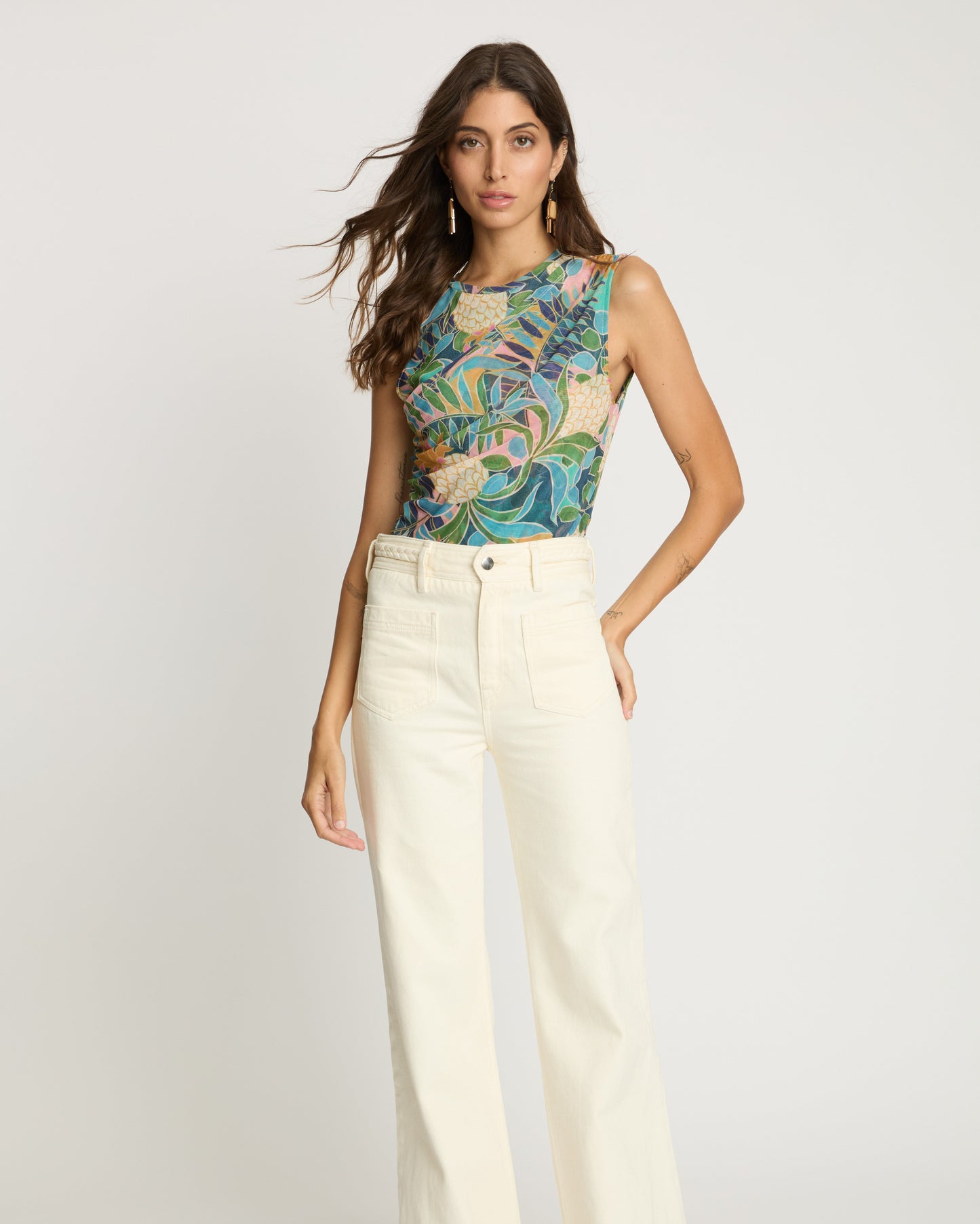 Woman wearing a colorful sleeveless top and white pants on a light gray background