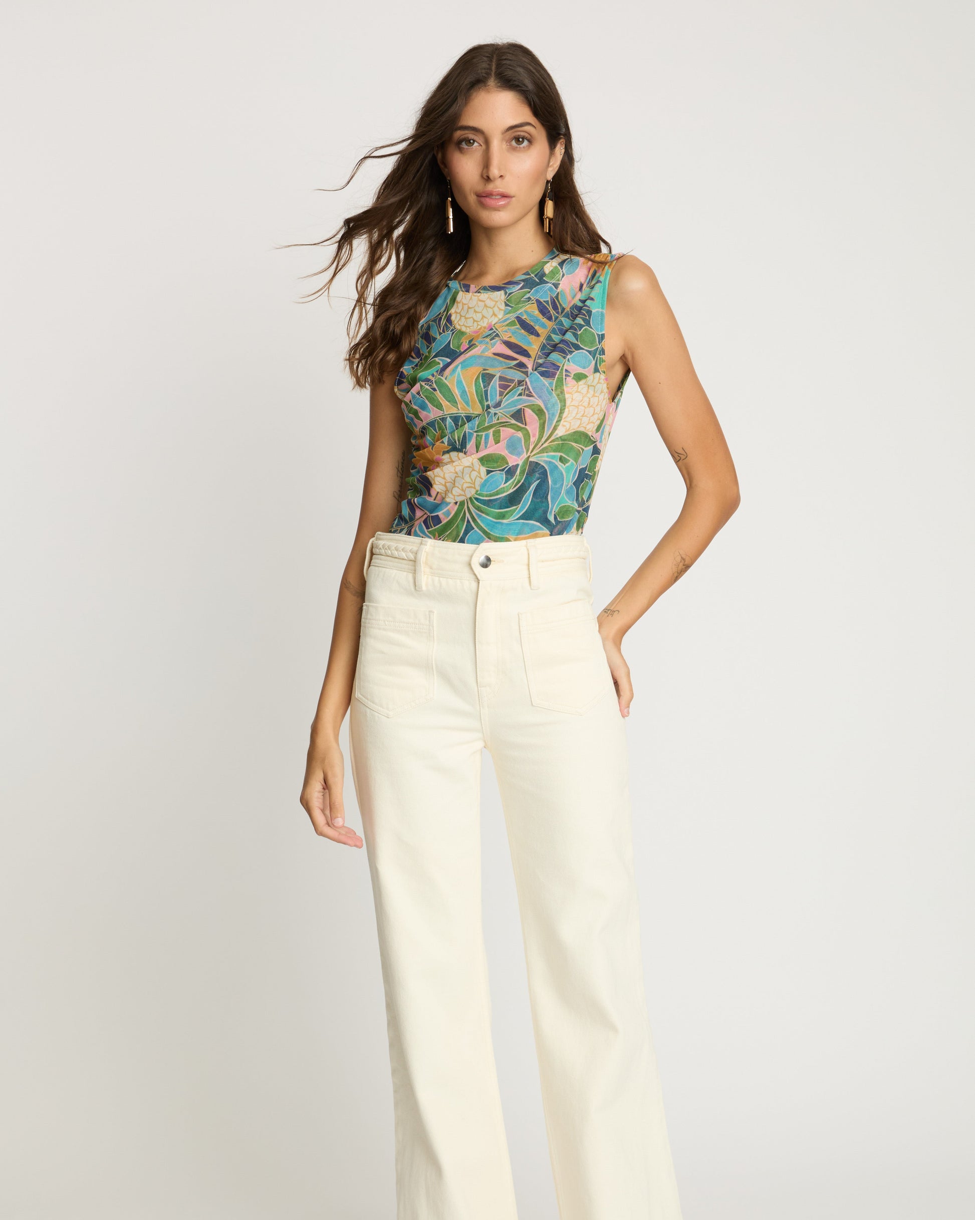 Woman wearing a colorful sleeveless top and white pants on a light gray background