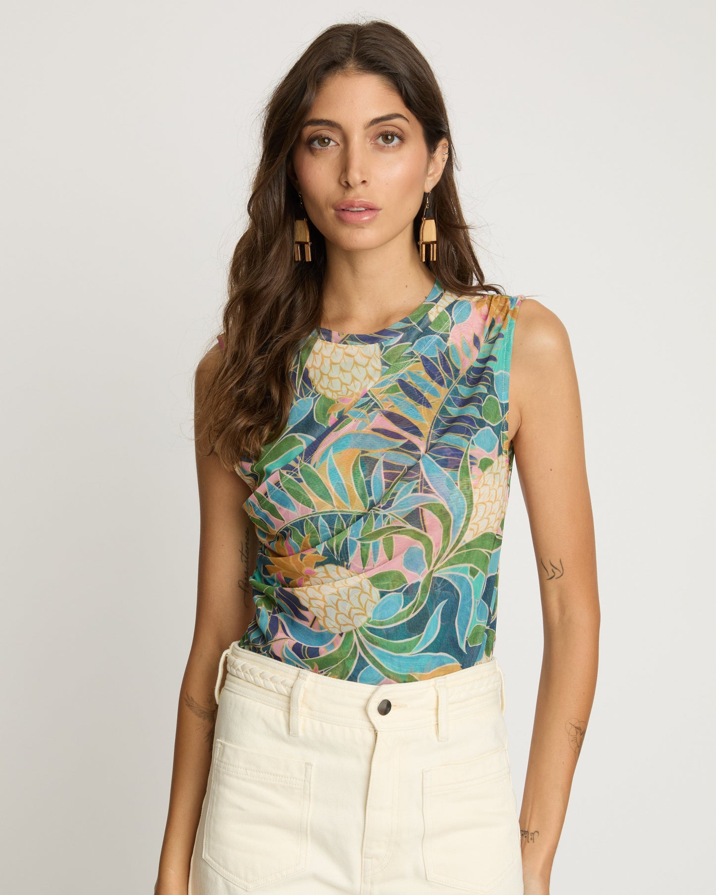 Woman wearing a colorful floral sleeveless top and white pants against a plain background