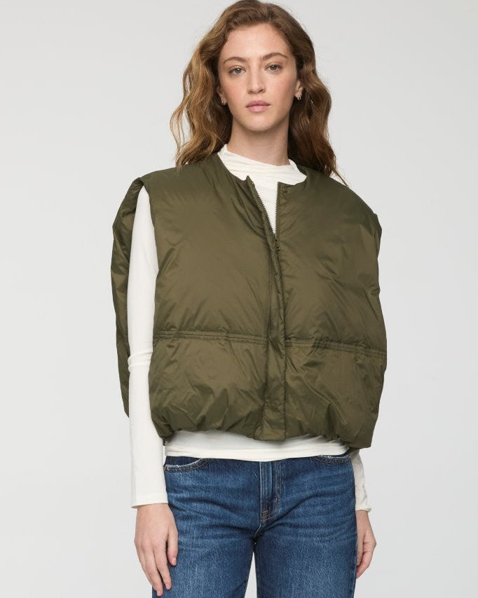 A woman modeling a sleeveless olive green quilted puffer vest over a white long-sleeve top and blue jeans, photographed against a light gray studio background.