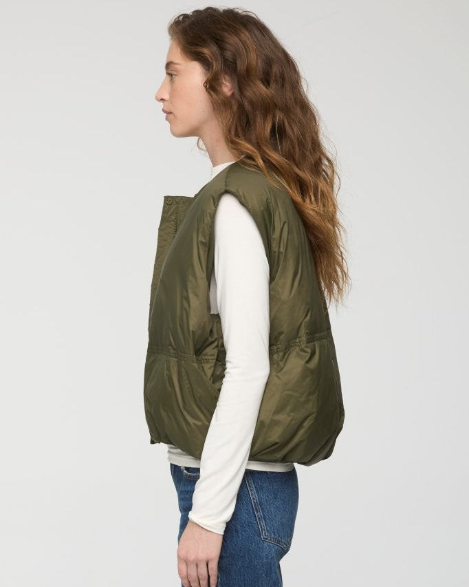 A woman shown in profile wearing an olive green quilted puffer vest over a white long-sleeve top and blue jeans, photographed against a plain light gray studio background.