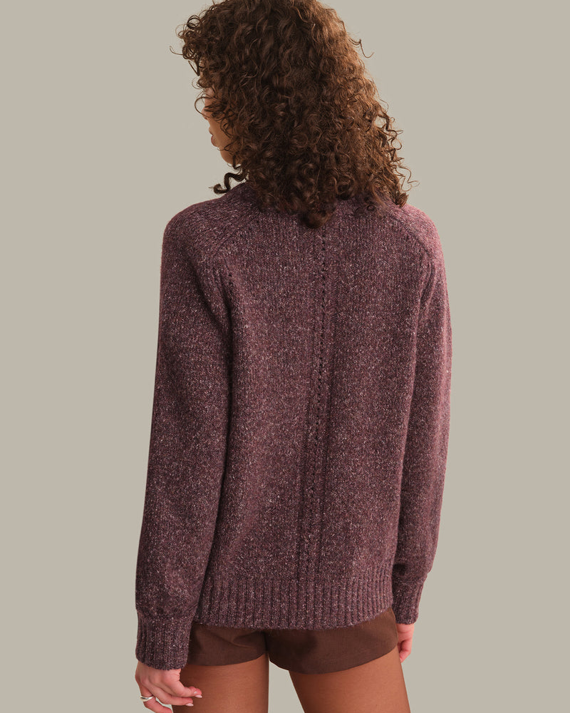 A woman with curly brown hair is shown from the back wearing a textured, heathered burgundy knit sweater and brown shorts against a solid light gray background.