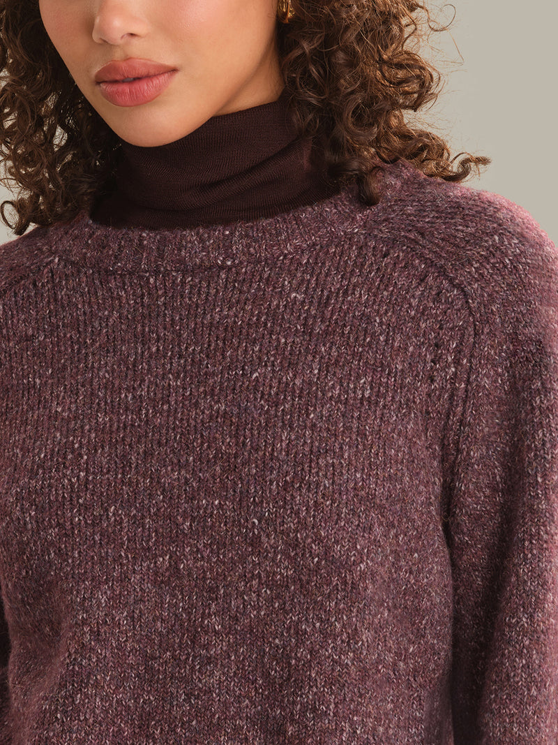 Close-up of a person wearing a ribbed knit turtleneck sweater in a heathered plum-brown color, with curly brown hair visible against a plain light beige background.