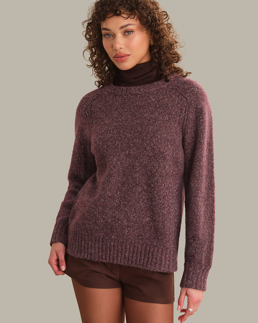 A woman wearing a heathered plum-purple knit sweater over a dark brown mock neck top and matching brown shorts, photographed against a solid light gray studio background.