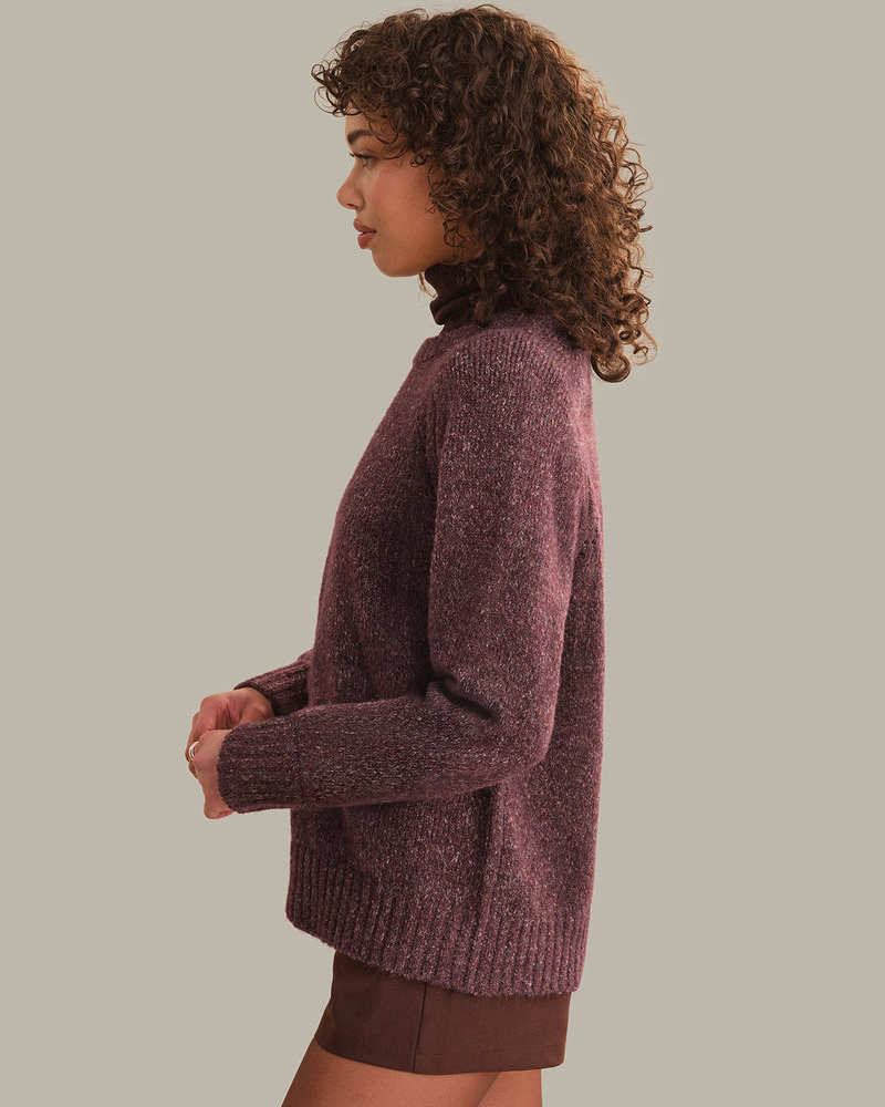 A woman shown in side profile wearing a fuzzy knit sweater in a muted plum-purple color, paired with brown shorts, against a solid light gray background.