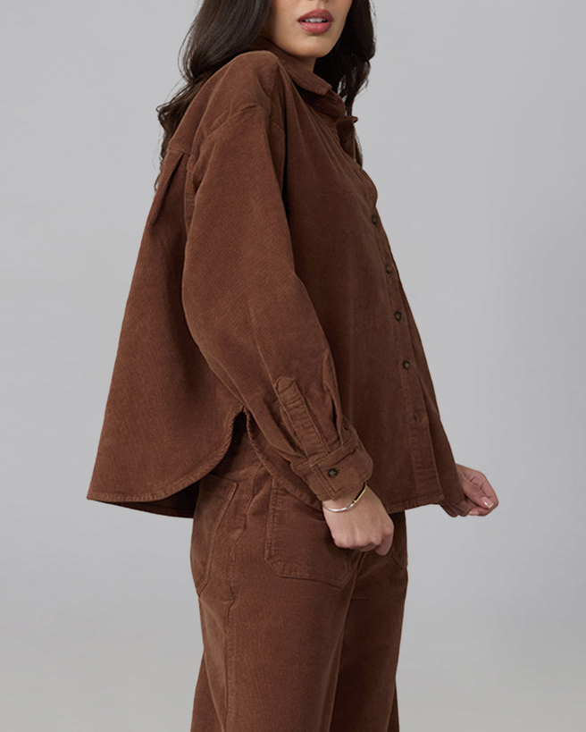 A woman wearing a brown corduroy button-front shirt jacket with matching brown corduroy trousers, shown from the side against a plain light gray studio background.