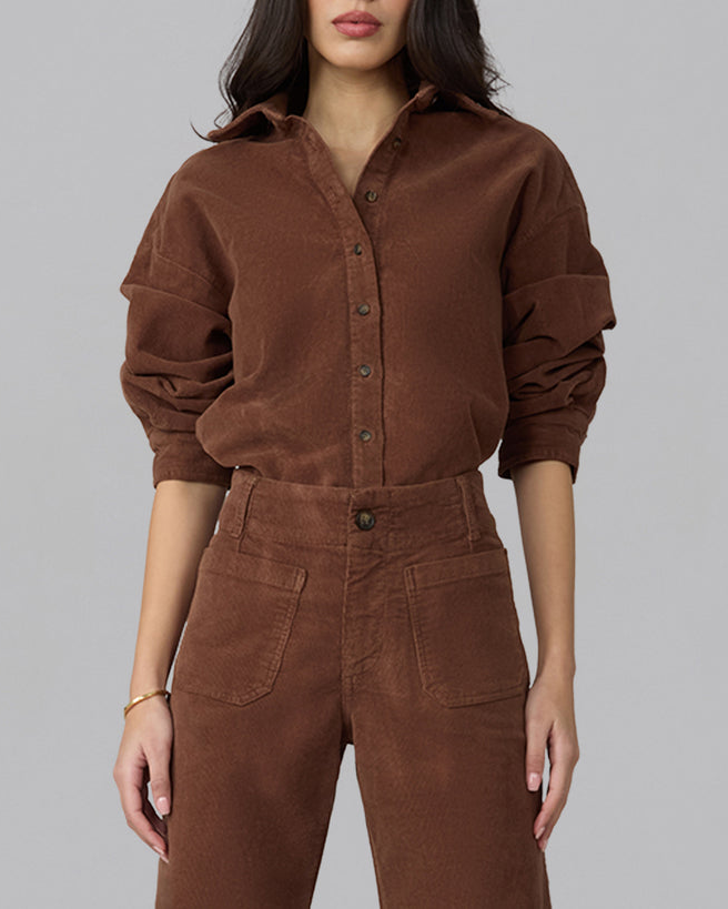 A woman wearing a brown corduroy button-up shirt and matching brown corduroy pants, shown from the mouth down against a solid gray studio background.