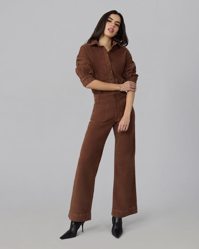 A woman modeling a brown corduroy jumpsuit with wide legs and a button-up front, paired with black pointed-toe ankle boots, against a solid light gray studio background.