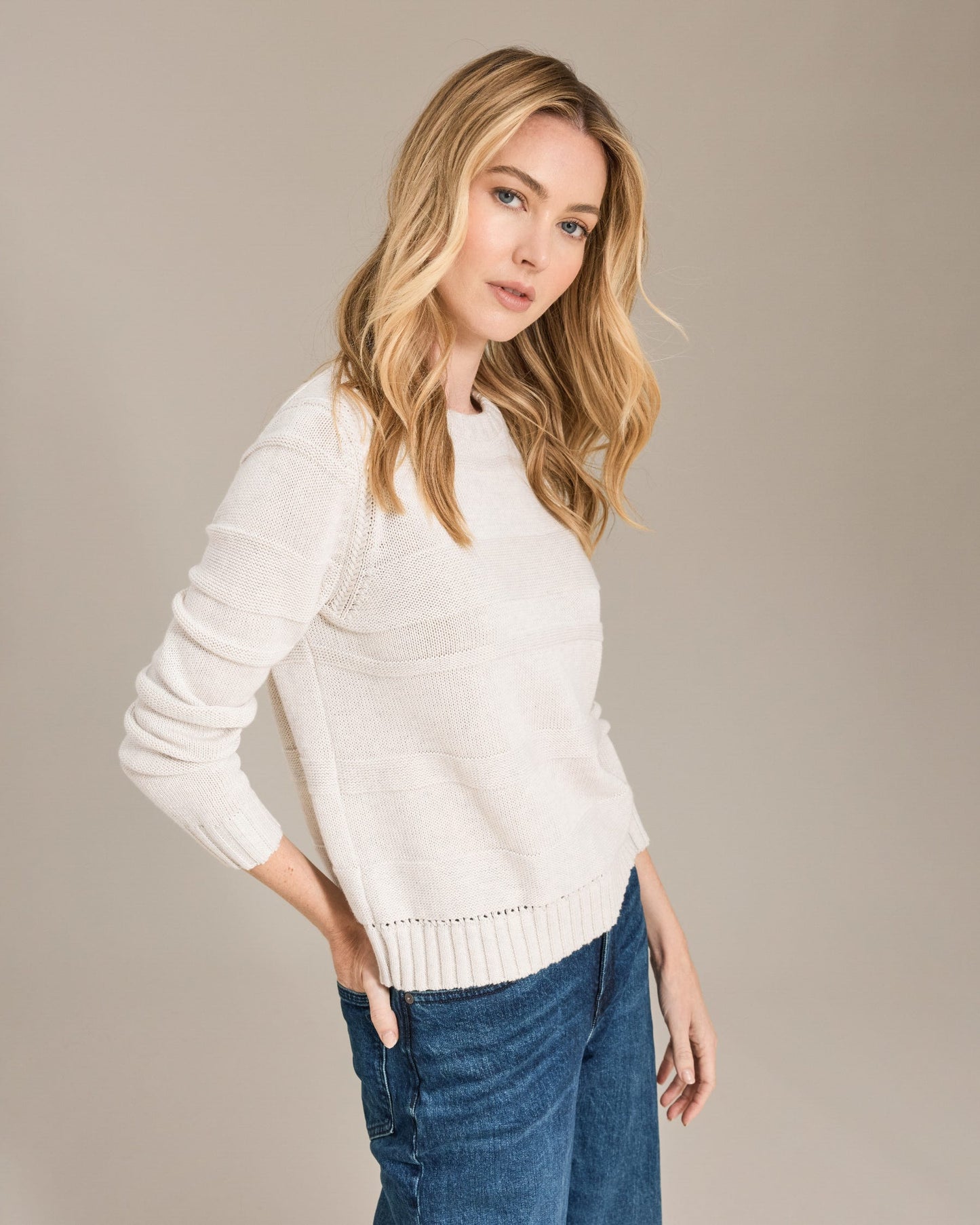 Woman wearing a white sweater and blue jeans against a beige background