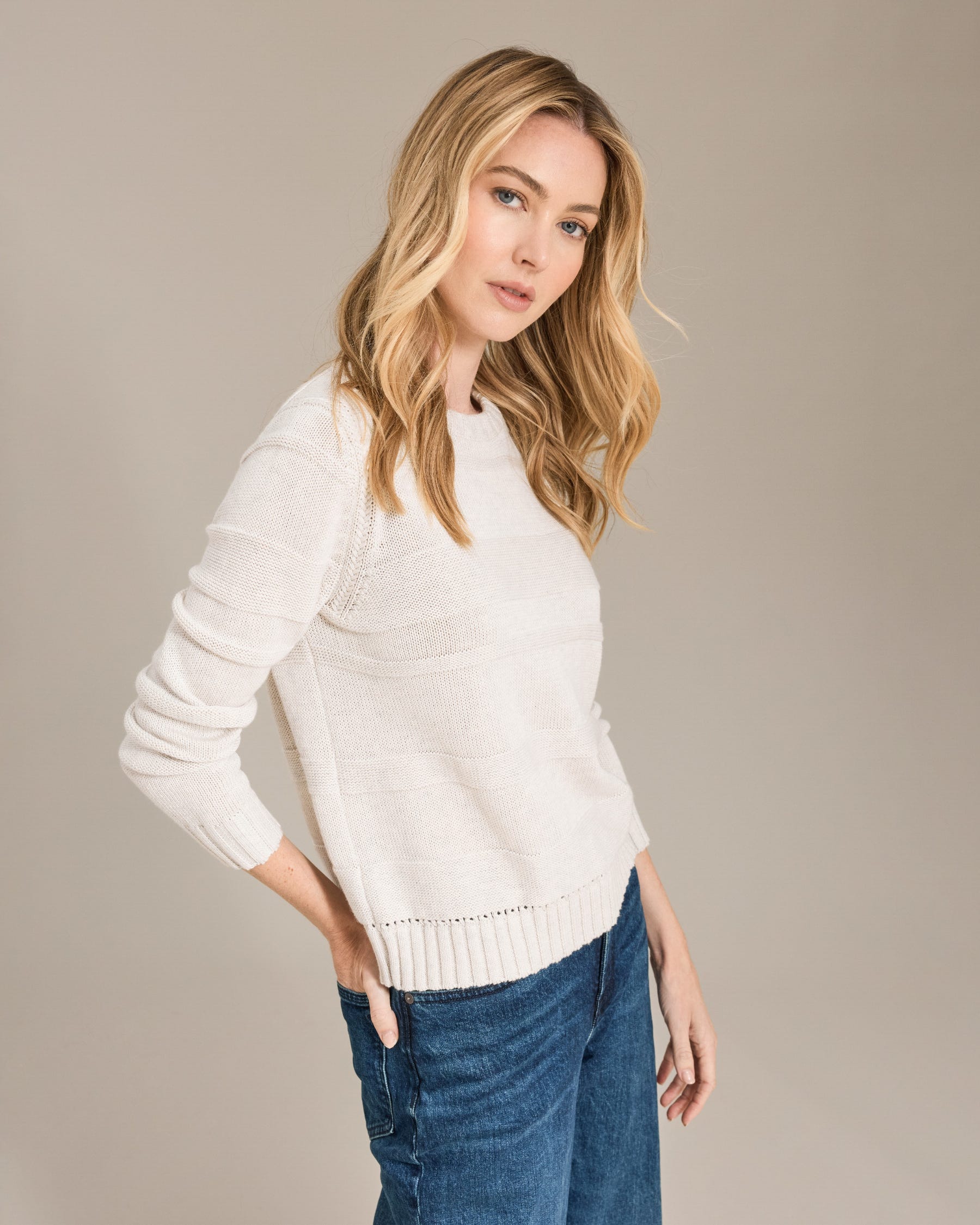 Woman wearing a white sweater and blue jeans against a beige background