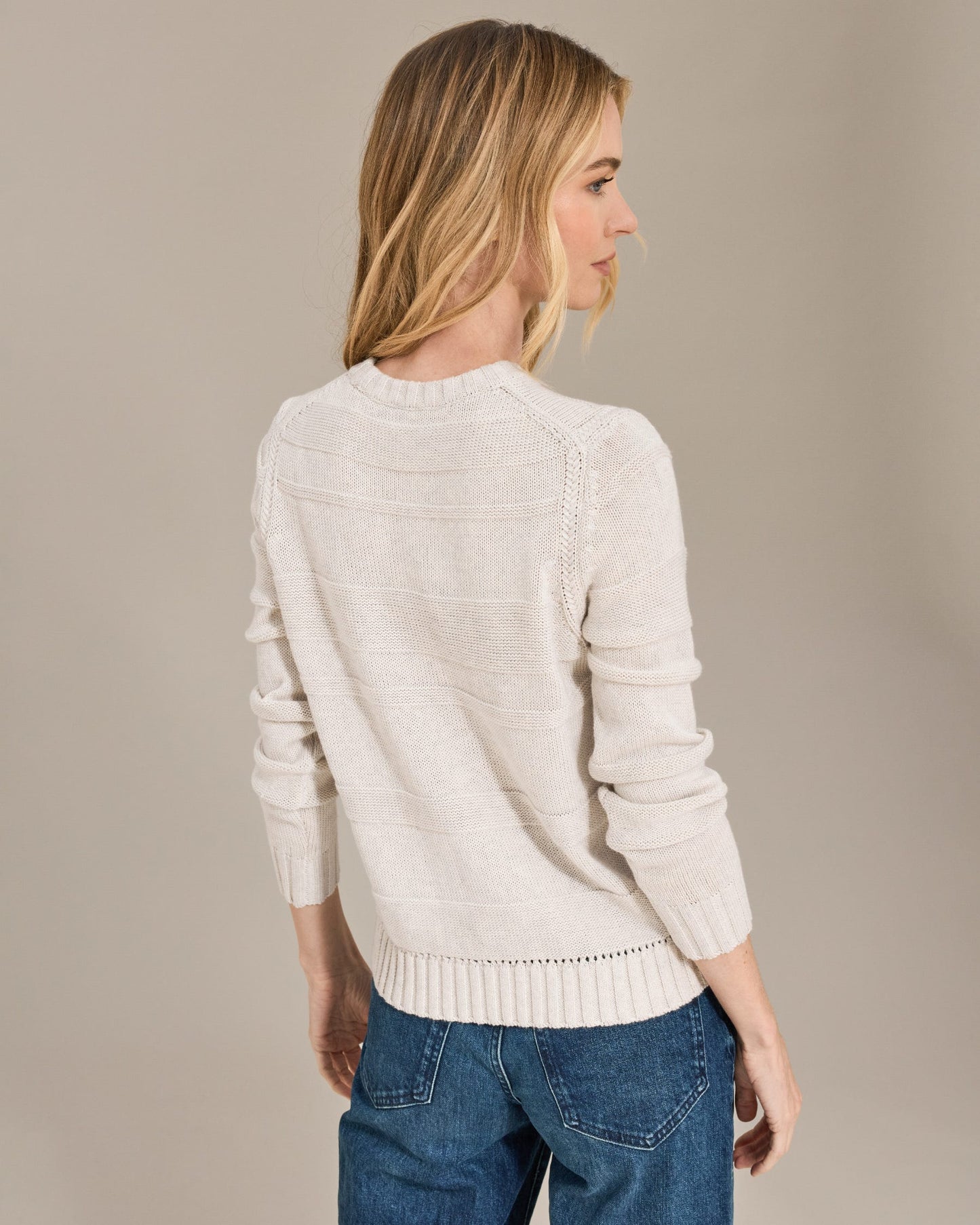 Woman wearing a light-colored sweater and blue jeans against a beige background