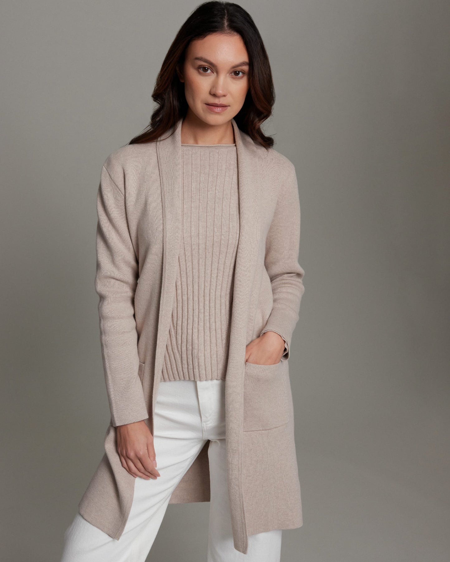 Woman wearing a beige cardigan over a sweater and white pants on a gray background