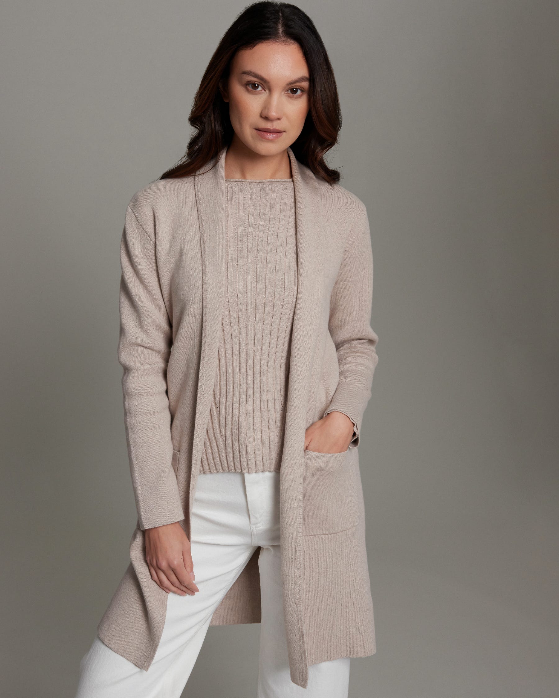 Woman wearing a beige cardigan over a sweater and white pants on a gray background