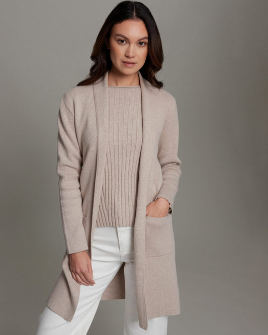 Woman wearing a beige cardigan over a sweater and white pants on a gray background