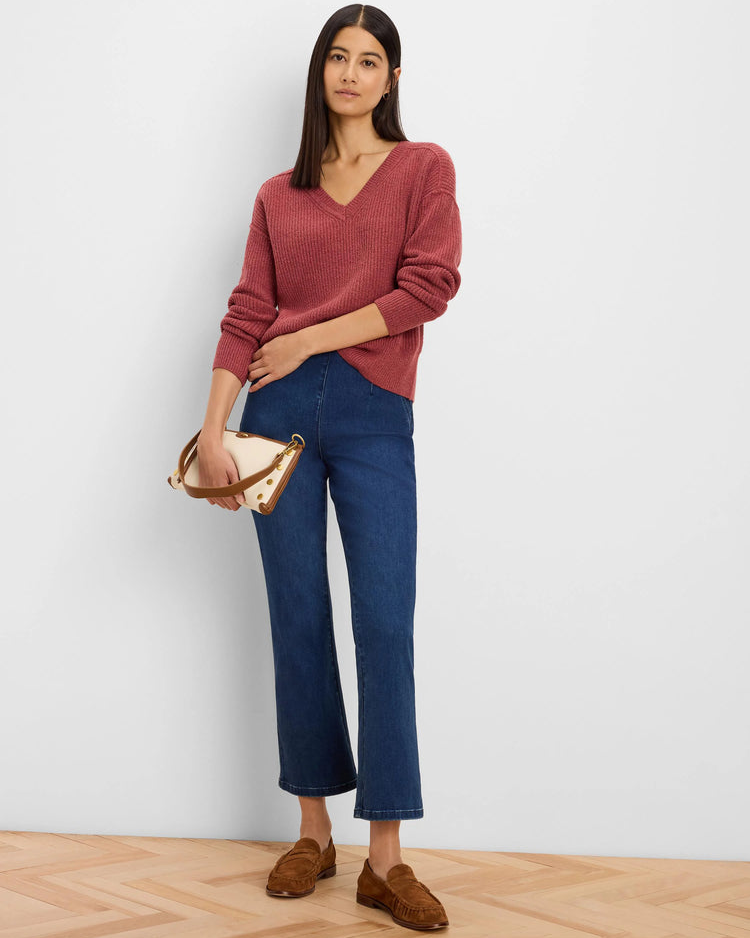 A woman stands against a light gray wall wearing a dusty rose ribbed V-neck sweater, dark blue cropped flare jeans, and brown suede loafers, while holding a beige handbag with brown trim.