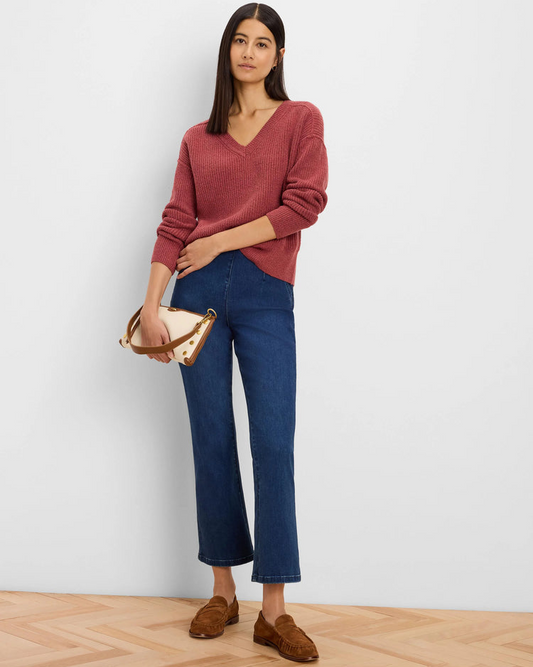 A woman stands against a light gray wall wearing a dusty rose ribbed V-neck sweater, dark blue cropped flare jeans, and brown suede loafers, while holding a beige handbag with brown trim.