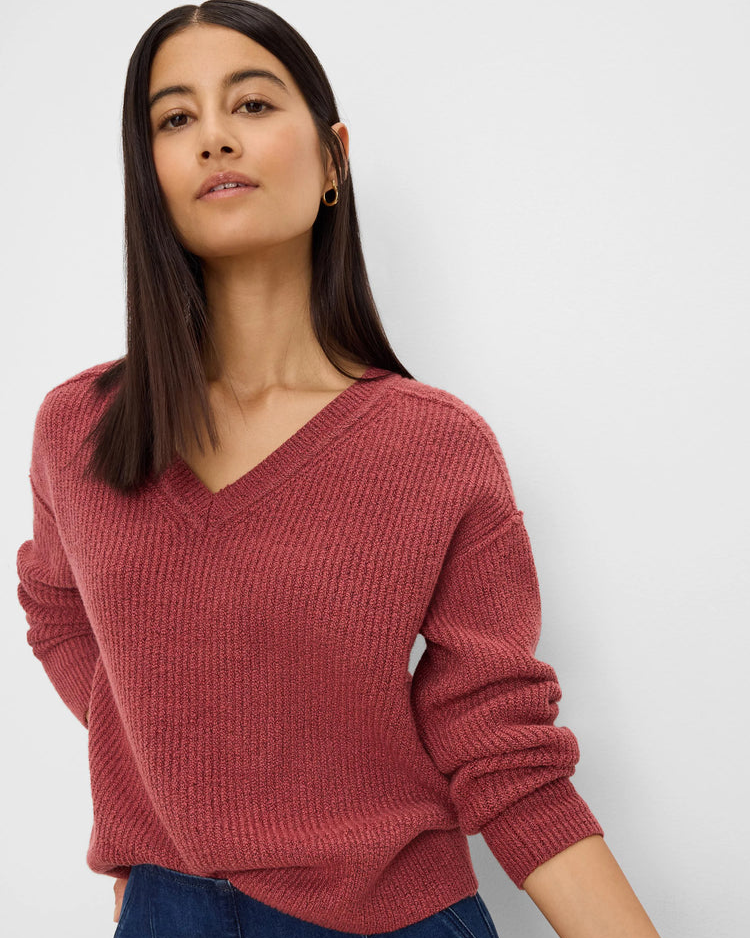 A woman wearing a ribbed knit V-neck sweater in a dusty rose color, paired with dark blue jeans, photographed against a light gray studio background.