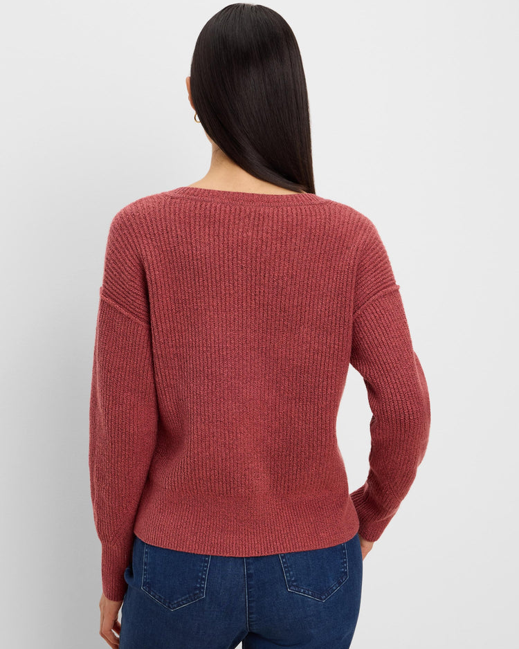 A woman viewed from the back wearing a ribbed knit sweater in a muted dusty rose color, paired with blue jeans, against a plain light gray studio background.