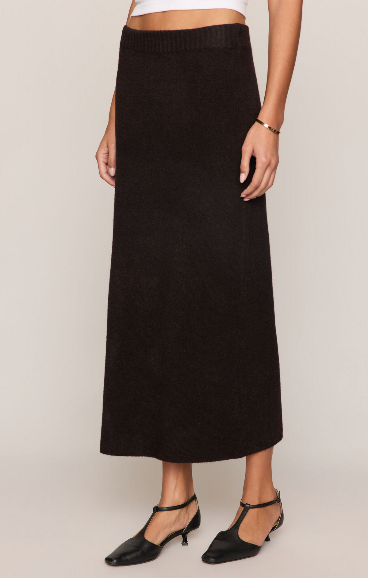 A model wearing a long, dark brown ribbed knit midi skirt with black T-strap kitten heels, shown from the waist down against a light beige studio background.