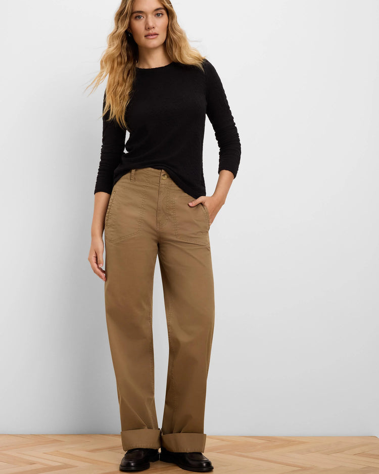 A woman modeling a black long-sleeve top and brown wide-leg trousers against a light gray wall and wood floor.
