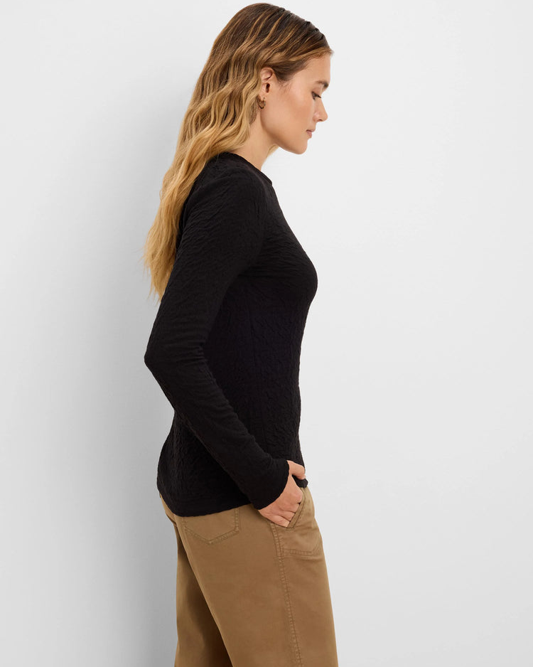 A woman shown in side profile wearing a fitted black long-sleeve top and tan pants against a plain light gray studio background.