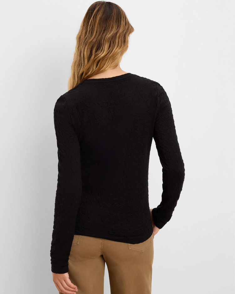 A woman shown from the back wearing a black long-sleeve knit top and tan pants against a light gray studio background.