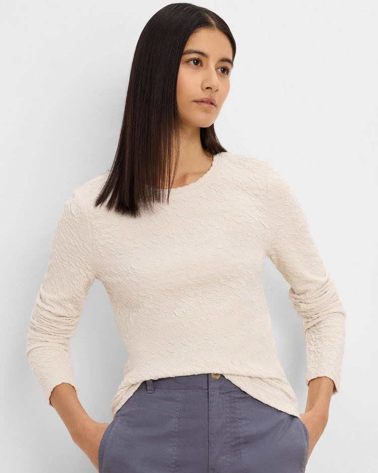 A woman wearing a textured cream long-sleeve crewneck top and slate blue trousers, standing against a light gray studio background.