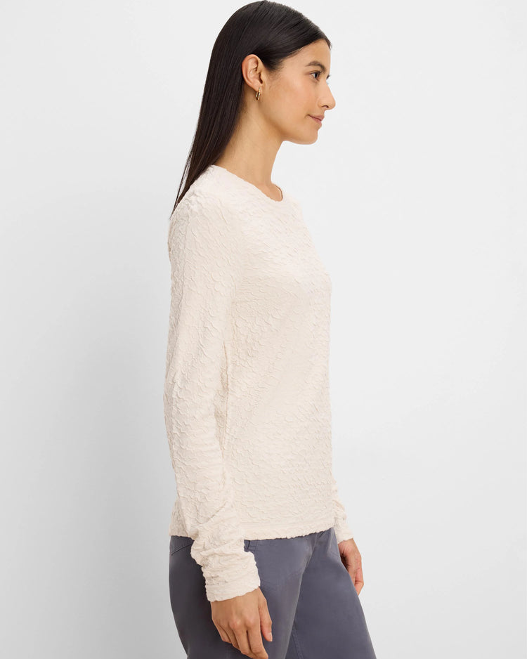 A woman shown in side profile wearing a textured, long-sleeve cream knit top and gray pants against a plain light gray background.