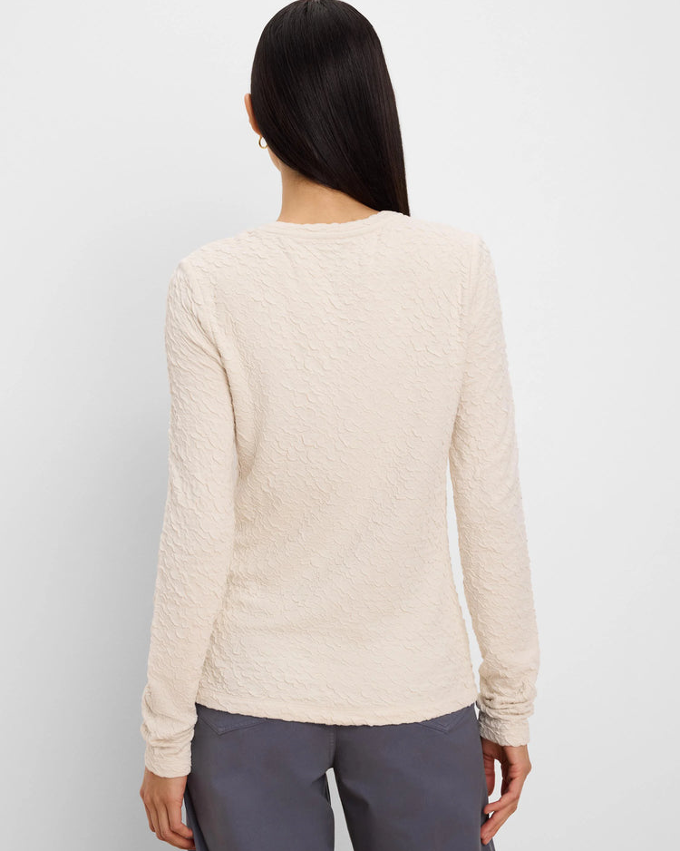 A woman viewed from the back wearing a textured, long-sleeve cream-colored top and dark gray pants against a plain light gray background.