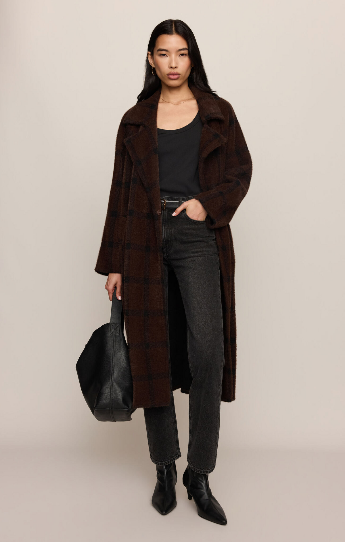A model stands against a plain beige studio background wearing a long brown plaid coat over a black top and dark gray jeans, holding a black tote bag.
