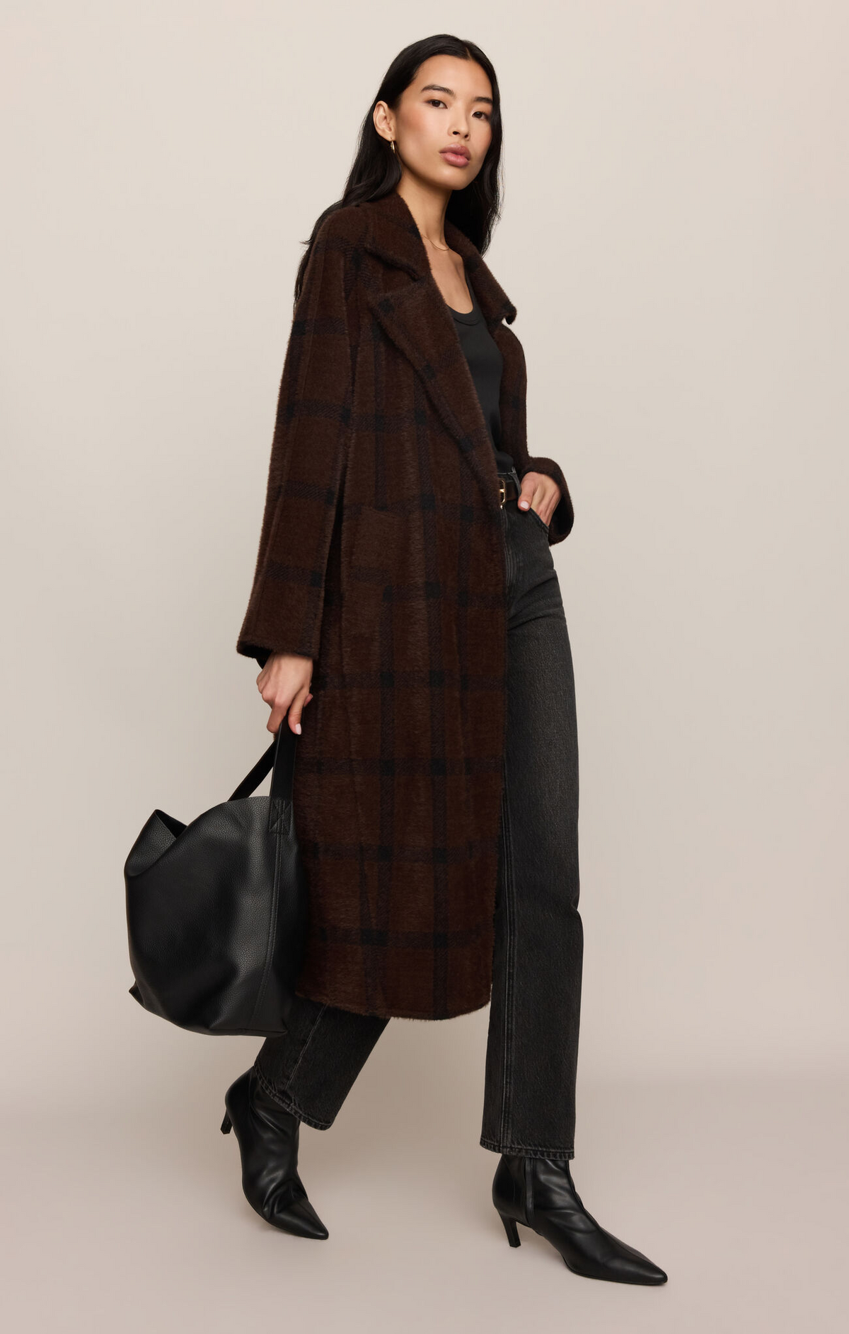 A model wearing a long brown plaid coat over a black outfit, holding a black tote bag against a solid beige studio background.