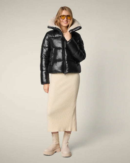 Woman wearing black puffer jacket with cream skirt against light grey background.