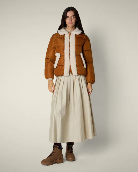 Woman wearing a brown puffer jacket with white fur lining, beige skirt, and brown boots on a plain background