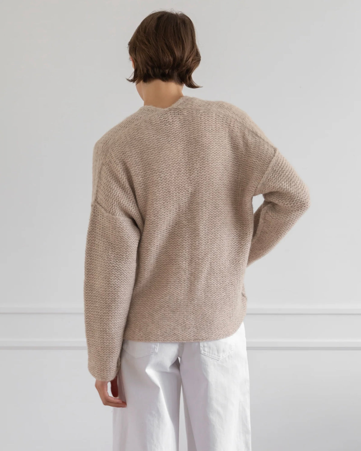 A person shown from the back wearing a textured knit sweater in a light taupe-beige color and white pants, standing against a plain white wall.