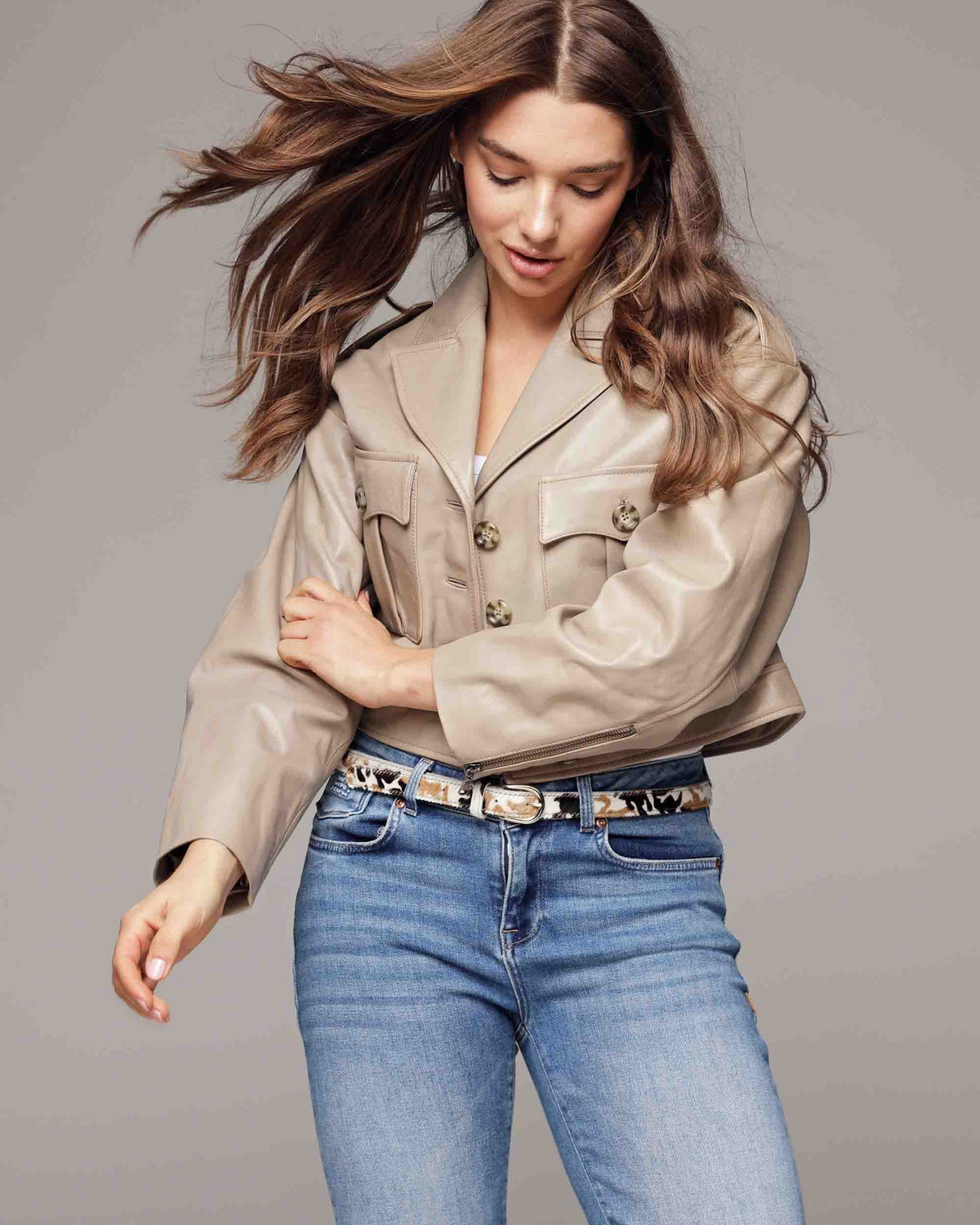 Woman wearing a beige jacket and blue jeans with a patterned belt against a gray background