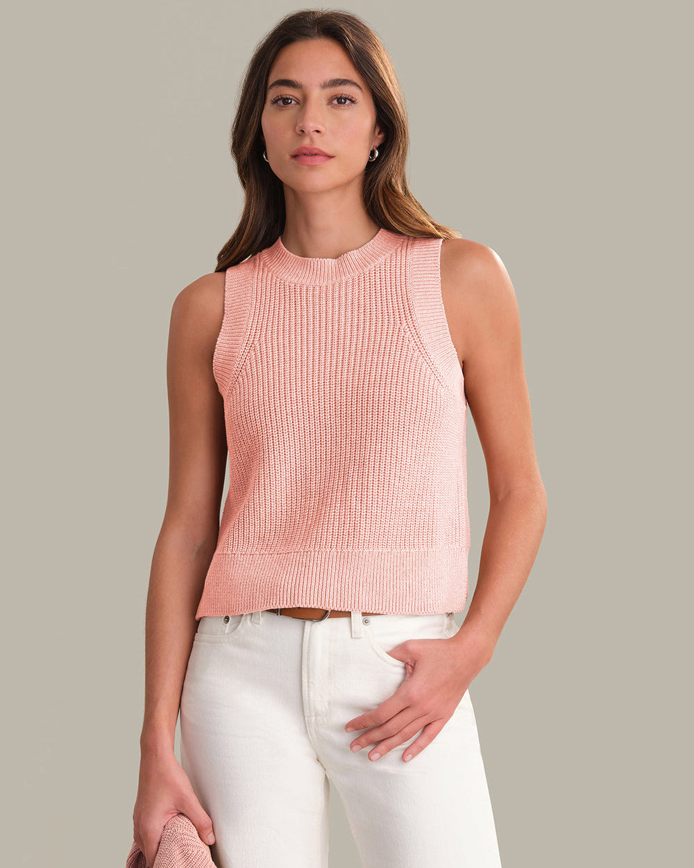 Woman wearing a pink sleeveless knitted top and white pants against a neutral background