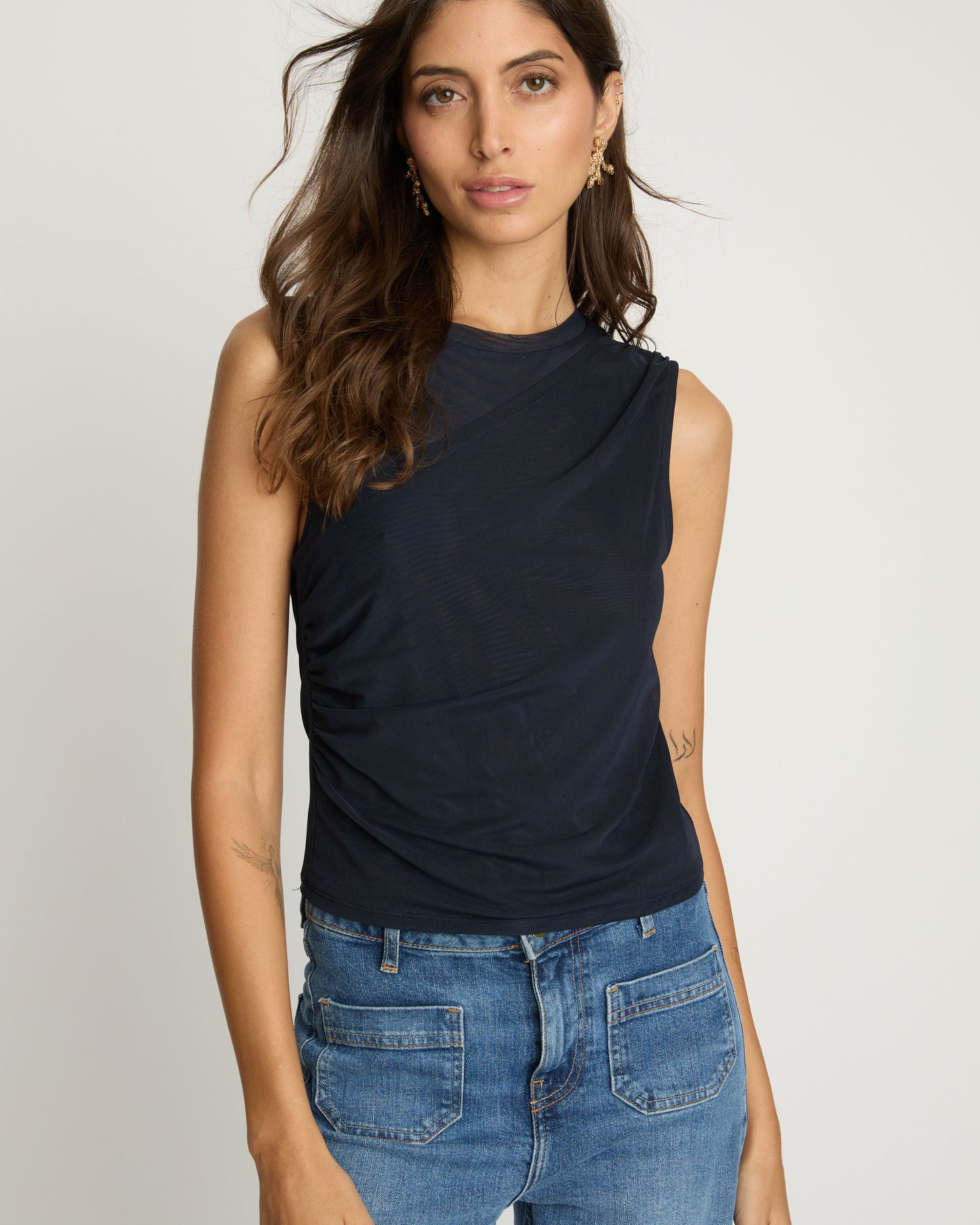 Woman wearing a navy sleeveless top and blue jeans on a plain background