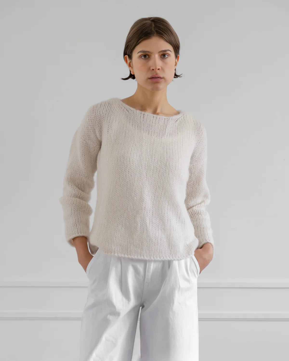 Woman wearing a beige sweater and white pants against a plain background
