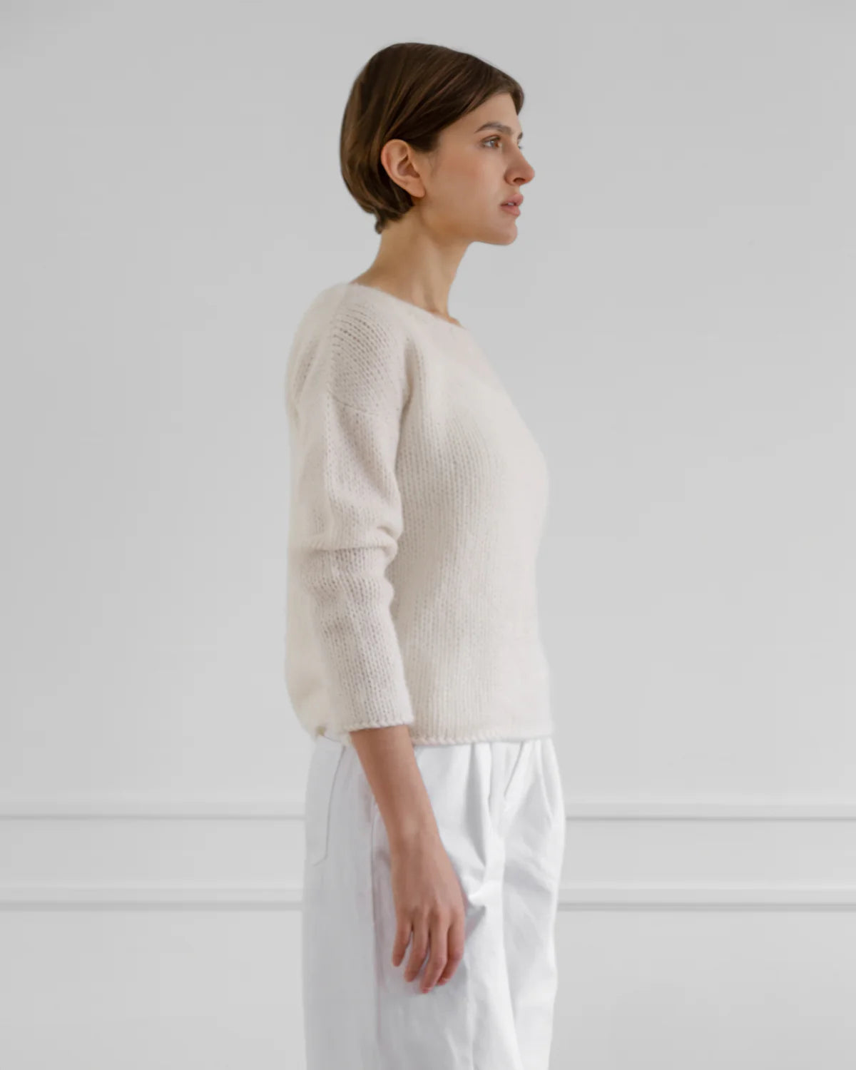 Woman wearing a beige sweater and white pants against a plain background