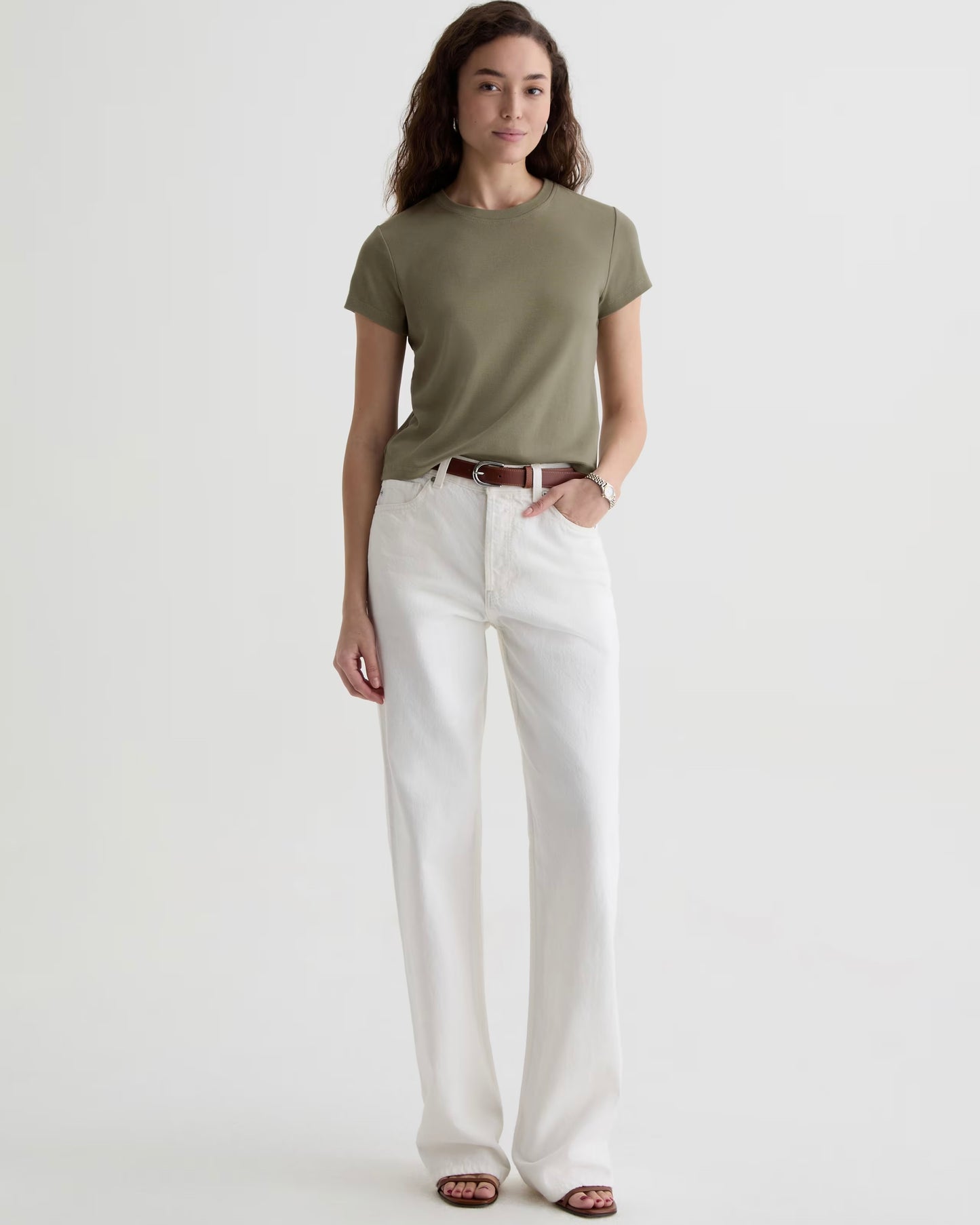 Woman wearing  green tee shirt with white jeans ad a brown belt standing in front of a white background