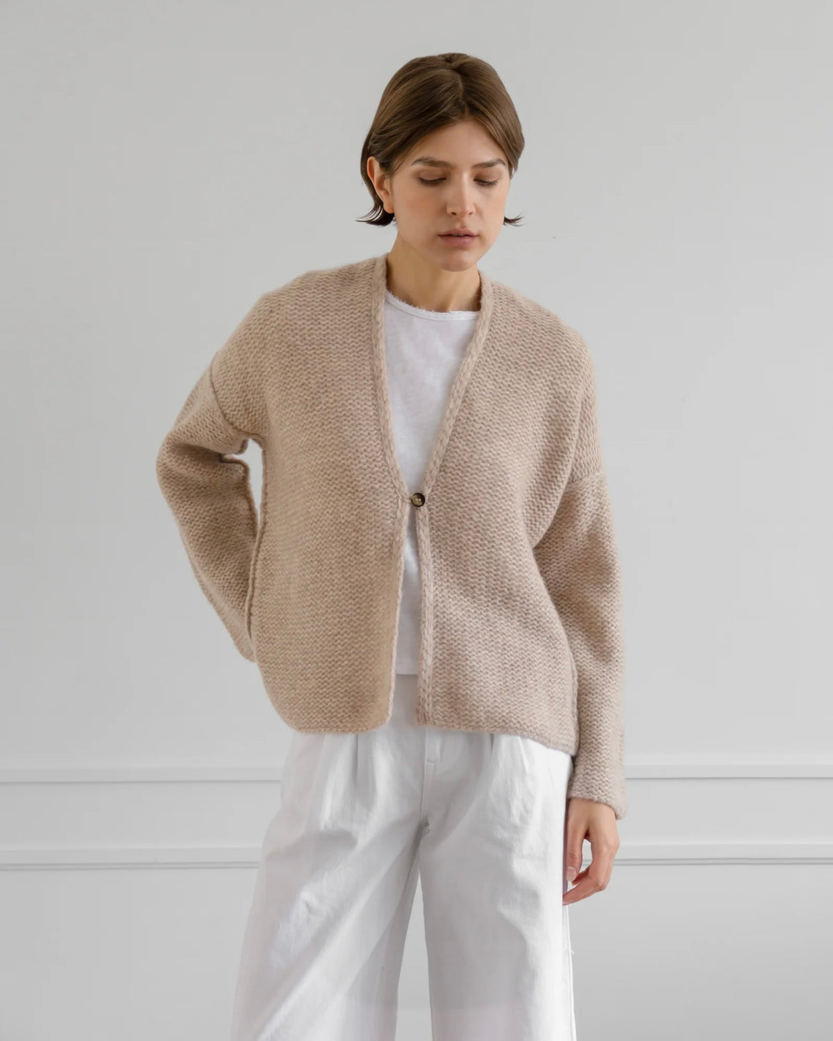 A woman wearing a beige textured knit cardigan over a white t-shirt and white trousers, standing against a light gray wall.